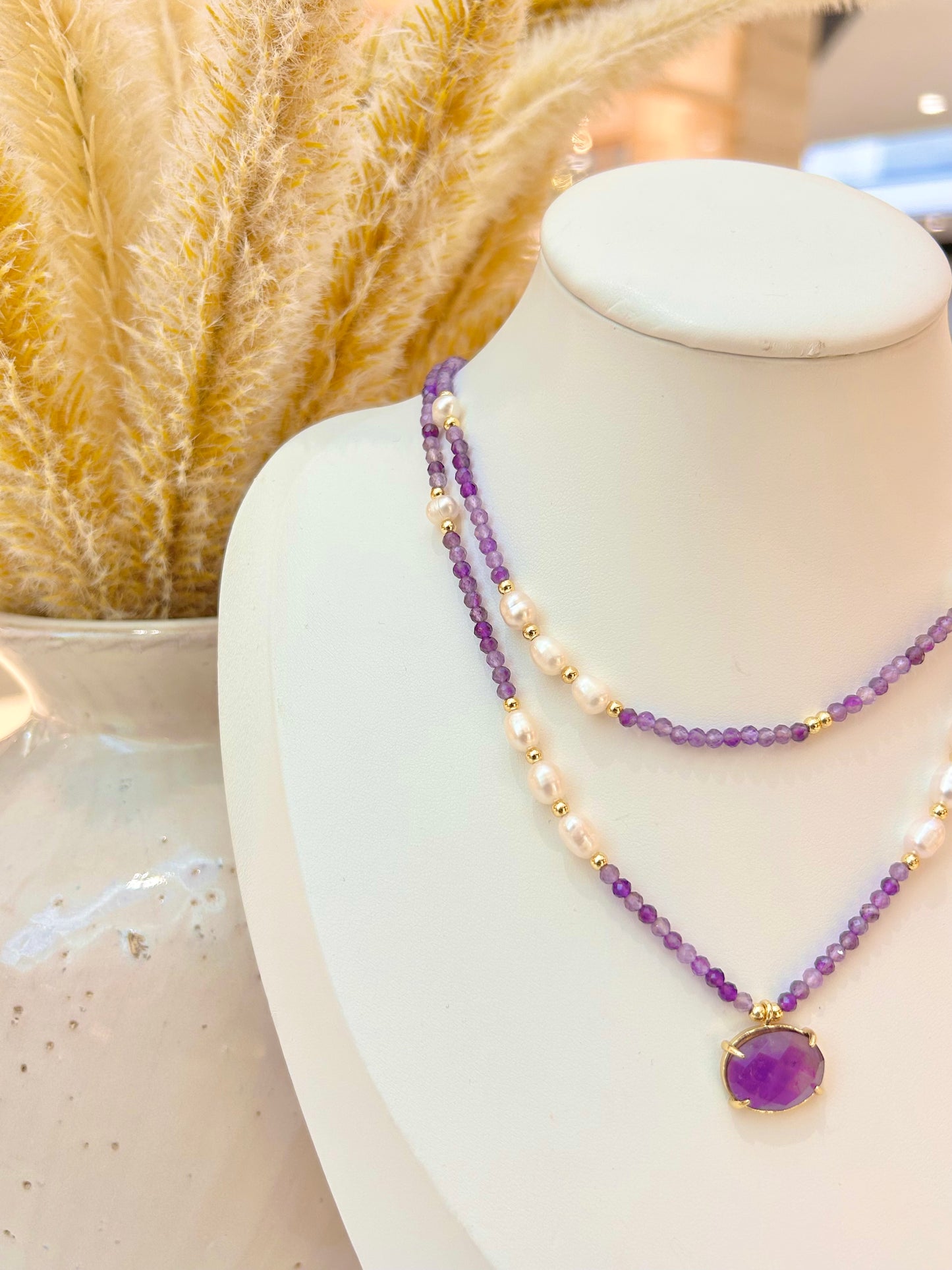 A double-strand necklace displayed on a white mannequin bust, featuring one strand of amethyst stone faceted beads and one strand of white pearls with small gold spacer beads. The necklace has a purple oval pendant in a gold bezel setting.