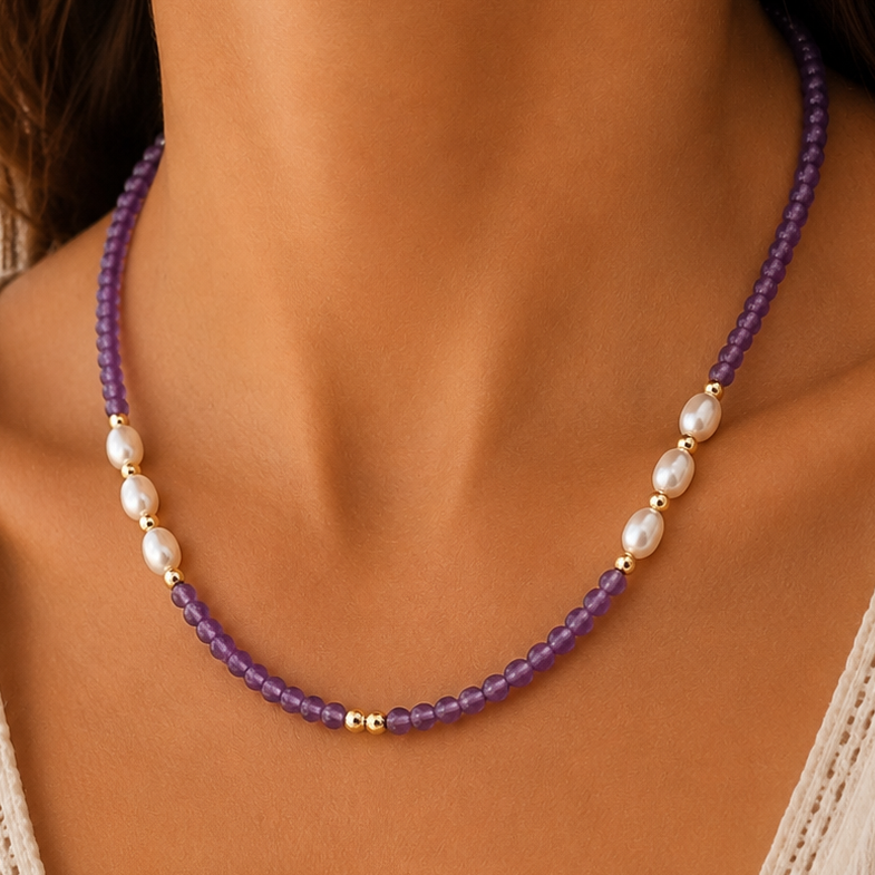 A close-up of a woman's neck and chest wearing a beaded necklace with amethyst beads, white pearl-like beads, and small gold accents, paired with a cream lace-trim V-neck top.