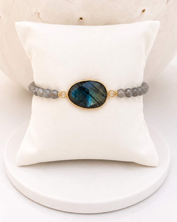 
                  
                    A beaded bracelet with grey labradorite stone beads and a central oval labradorite stone in a gold bezel setting, displayed on a white cushion.
                  
                