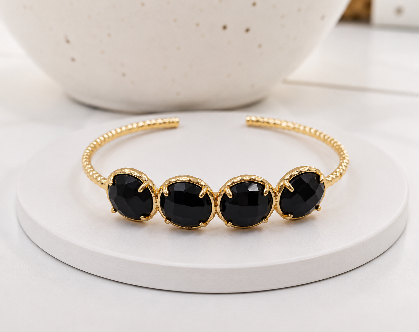 A gold-tone open cuff bracelet featuring four large, faceted black obsidian gemstones, displayed on a white round pedestal with a soft-focus ceramic bowl in the background.