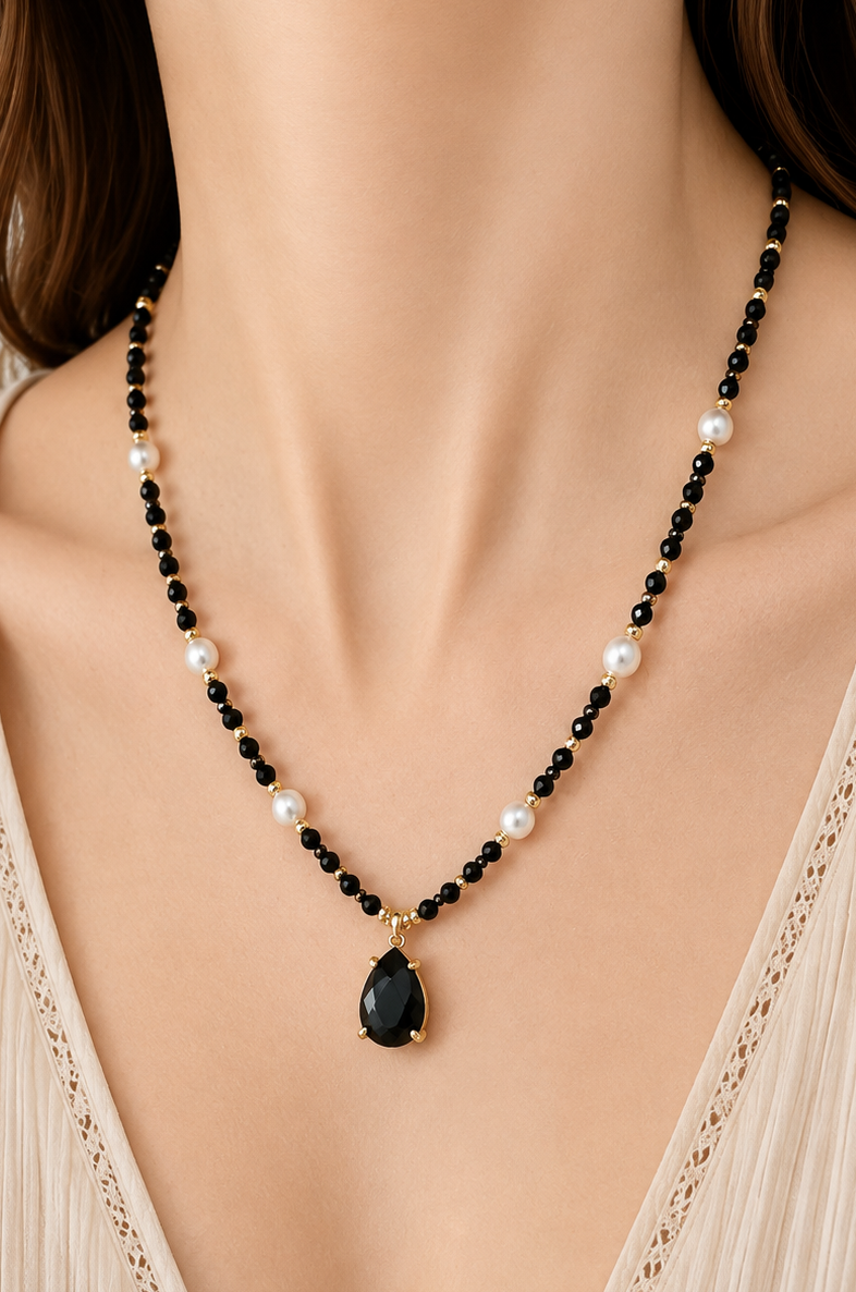A close-up of a woman wearing a beaded necklace with black obsidian and white pearl beads and a teardrop-shaped black obsidian pendant with gold detailing, paired with a beige lace-trimmed top.