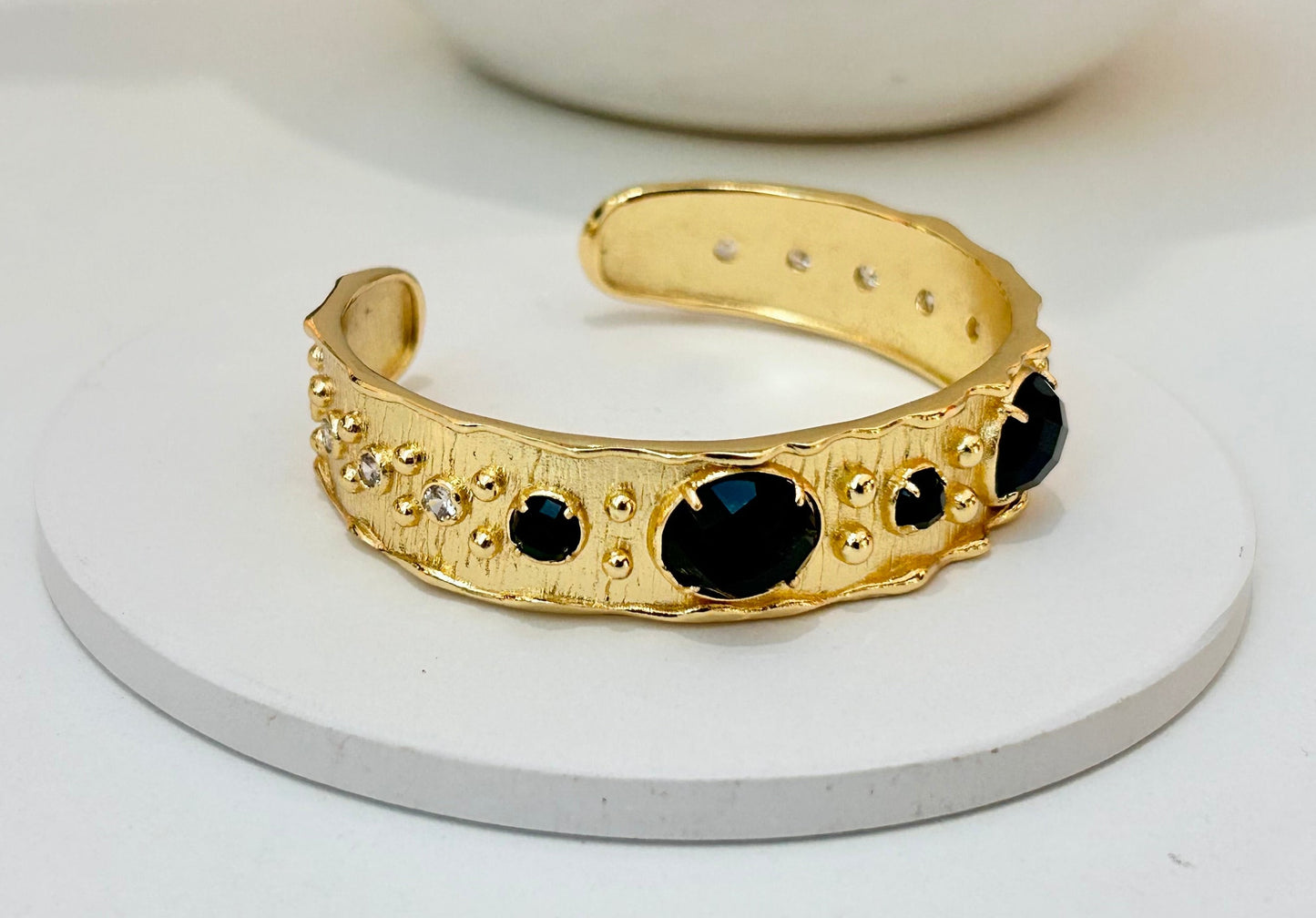 
                  
                    A textured gold cuff bracelet with black obsidian and round stones, displayed on a round white coaster.
                  
                