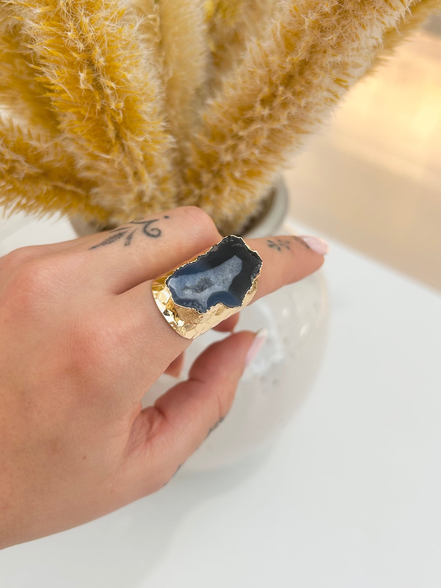A hand wearing a chunky ring made of a polished dark blue geode slice set in textured gold-toned metal, with dried pampas grass in the background.