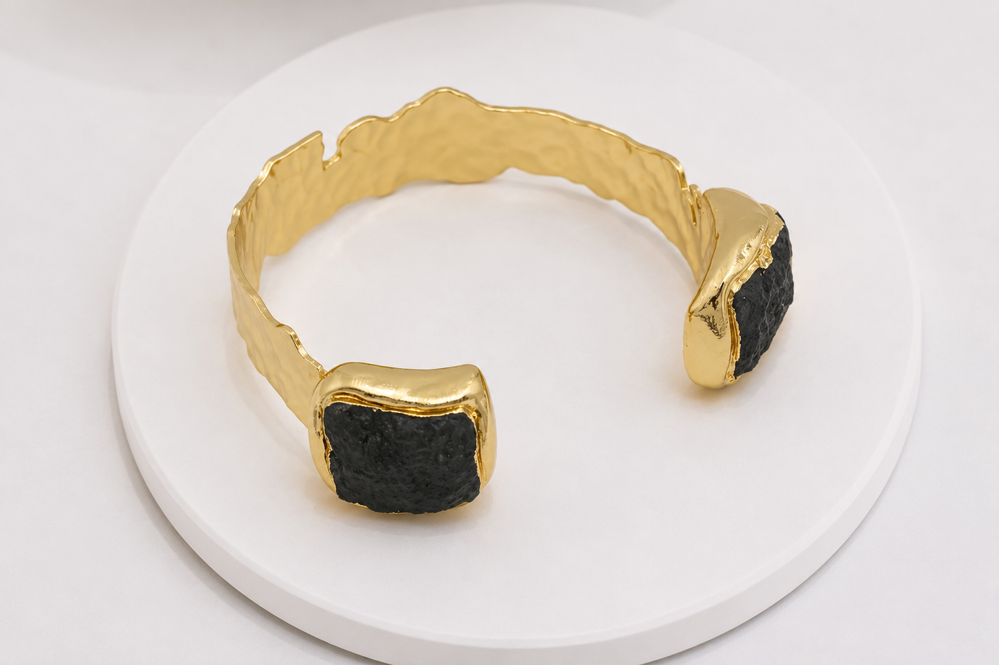 A gold cuff bracelet with two rough black stone inlays, displayed on a white circular pedestal.