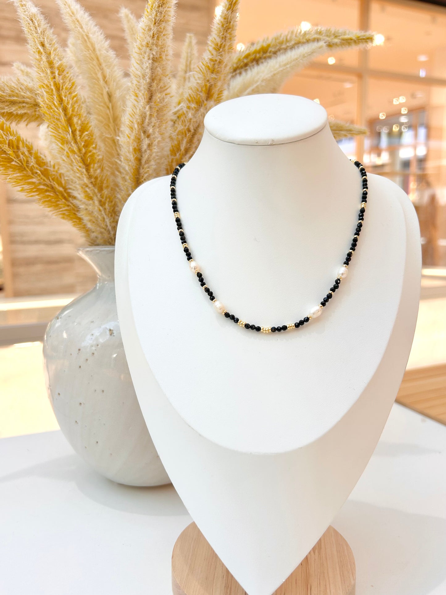 
                  
                    A beaded necklace with black obsidian and freshwater pearl beads displayed on a white jewellery bust, with dried pampas grass in a vase in the background.
                  
                