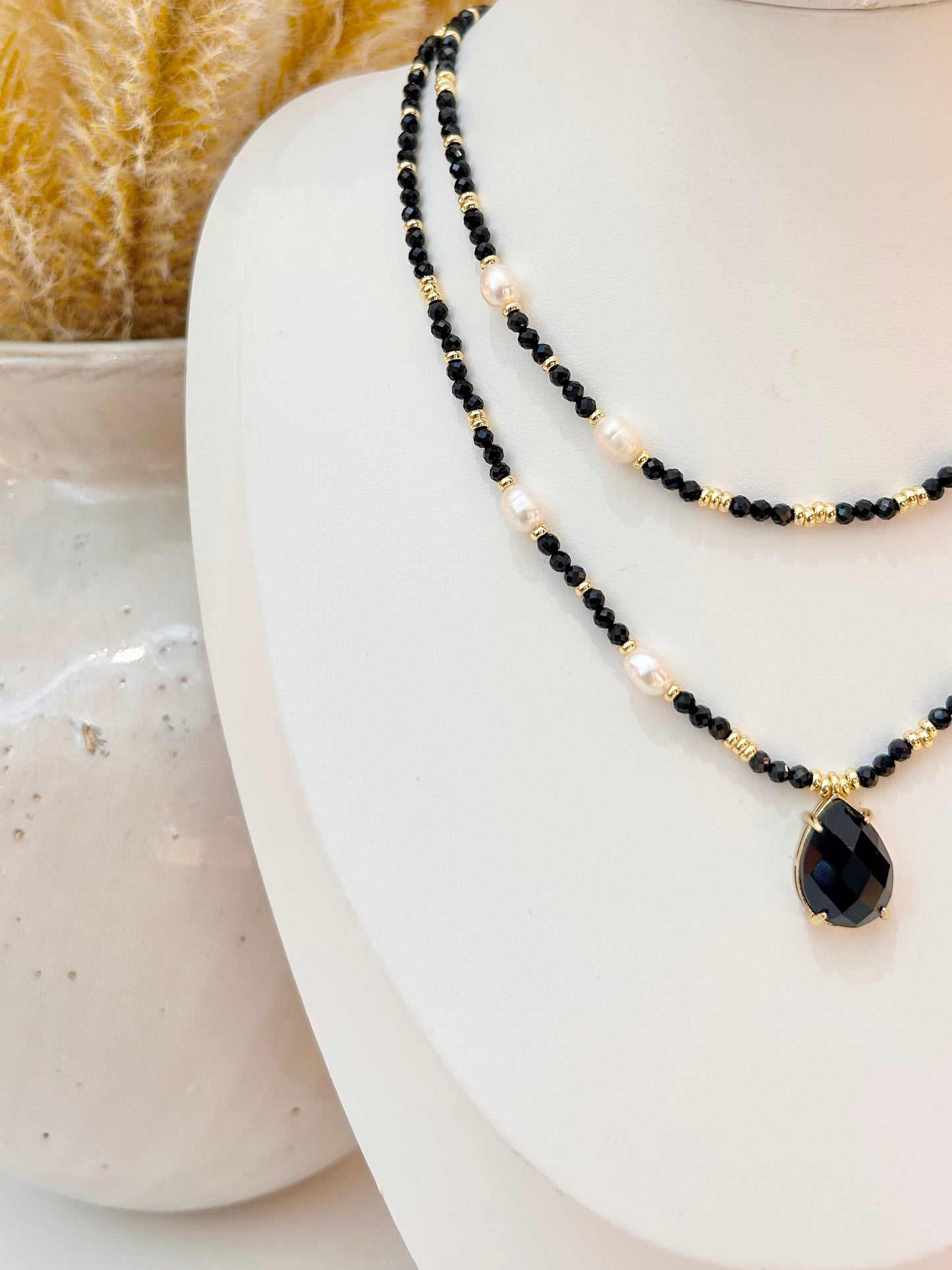 A double-strand beaded necklace displayed on a white mannequin bust. The necklace features black obsidian beads, small gold spacer beads, and white pearl-like beads, with a teardrop-shaped black stone pendant set in gold at the bottom right.
