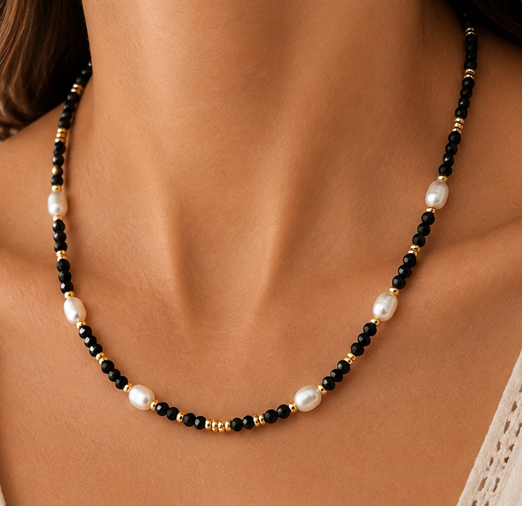 A close-up of a woman wearing a delicate beaded necklace with white pearls and black obsidian beads separated by small gold spacer beads, paired with a cream-colored eyelet top.