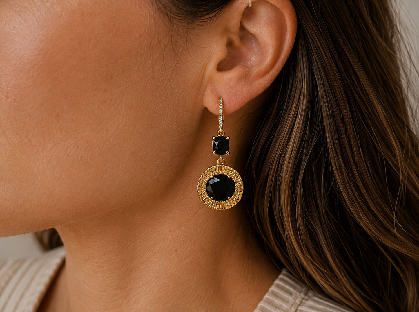A close-up of a woman's ear wearing a gold drop earring with a large round black obsidian gemstone and a smaller square black obsidian gemstone above it.