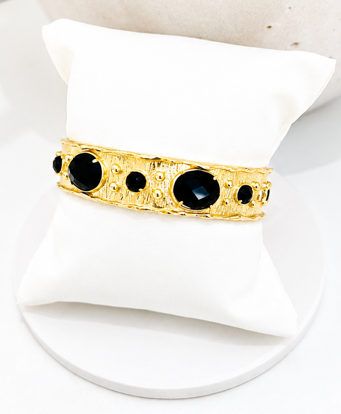 
                  
                    A gold bangle bracelet with black obsidian gemstones, displayed on a white cushion stand.
                  
                