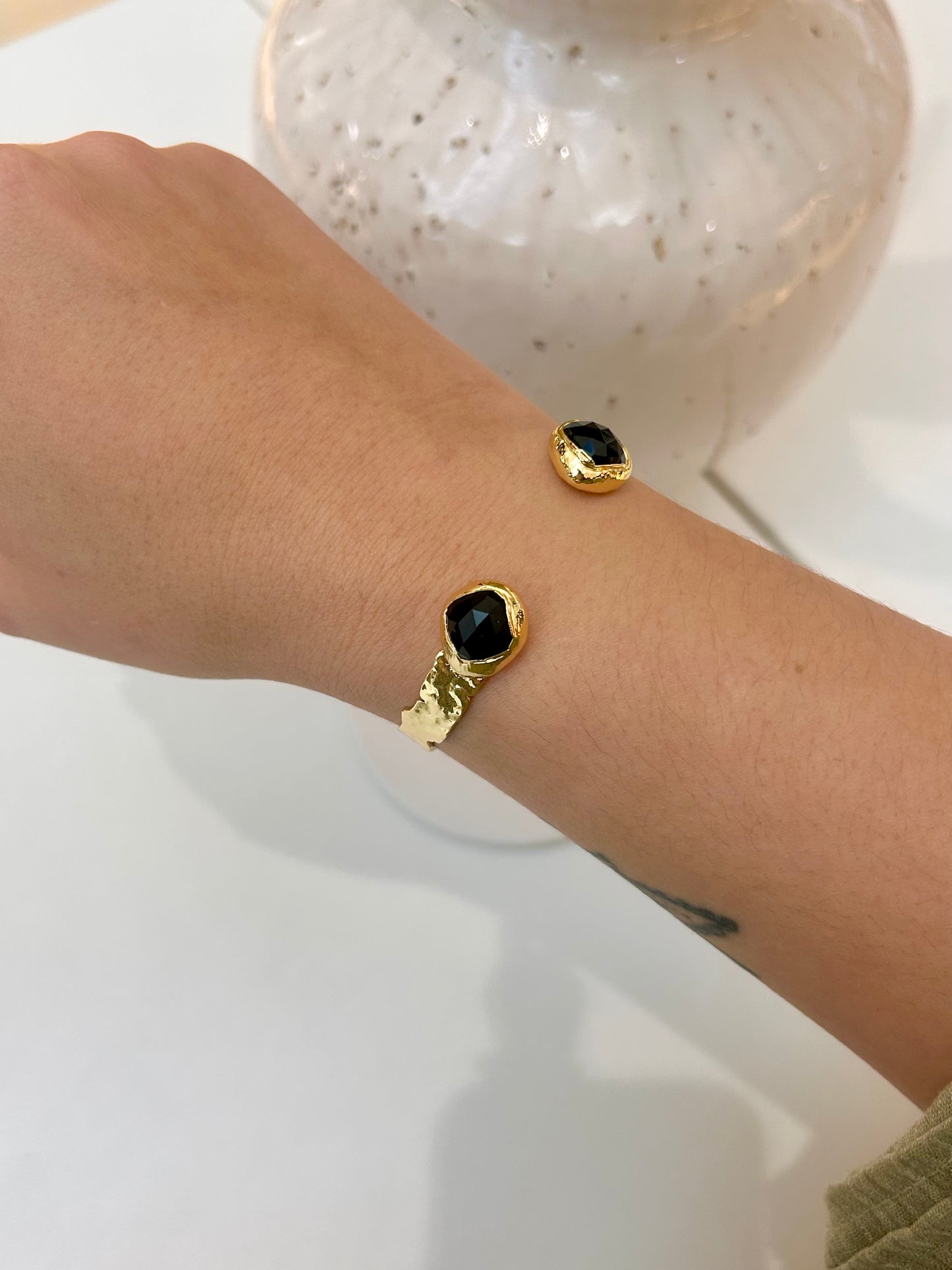 A close-up of a person's wrist wearing a gold bangle, each with a round black obsidian stone, against a light background with a decorative vase.