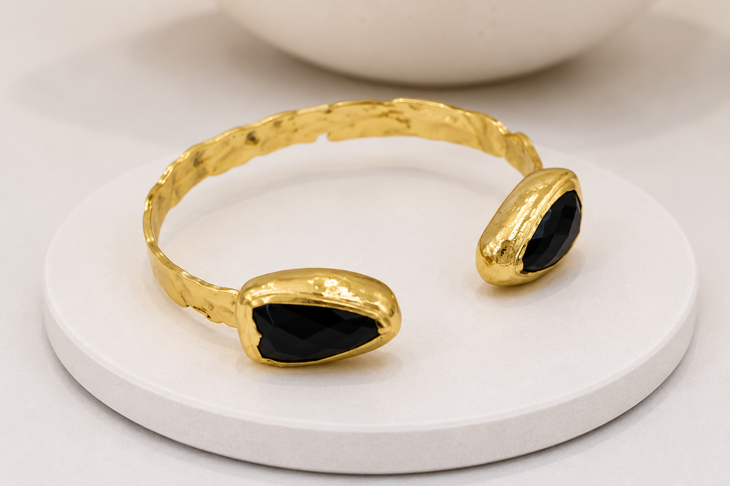 A gold-toned open cuff bracelet with two teardrop-shaped black obsidian accents, displayed on a round white pedestal.