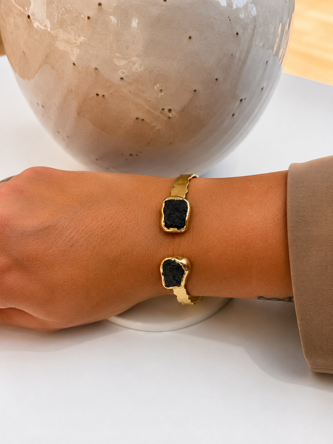 A close-up of a person's wrist wearing a gold-tone double cuff bracelet with irregular dark blue-grey natural stone inlays, resting on a white surface with a ceramic vase in the background.