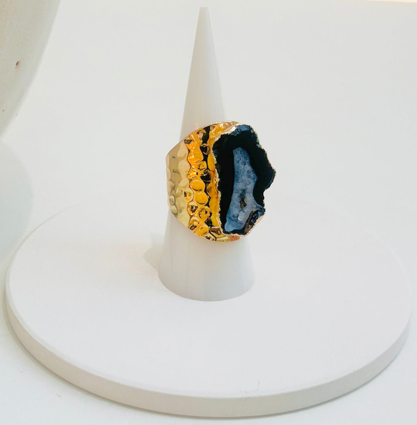 
                  
                    A statement ring featuring a split geode design, with one half in textured gold and the other in a dark blue and black agate geode slice, displayed on a white cone stand.
                  
                
