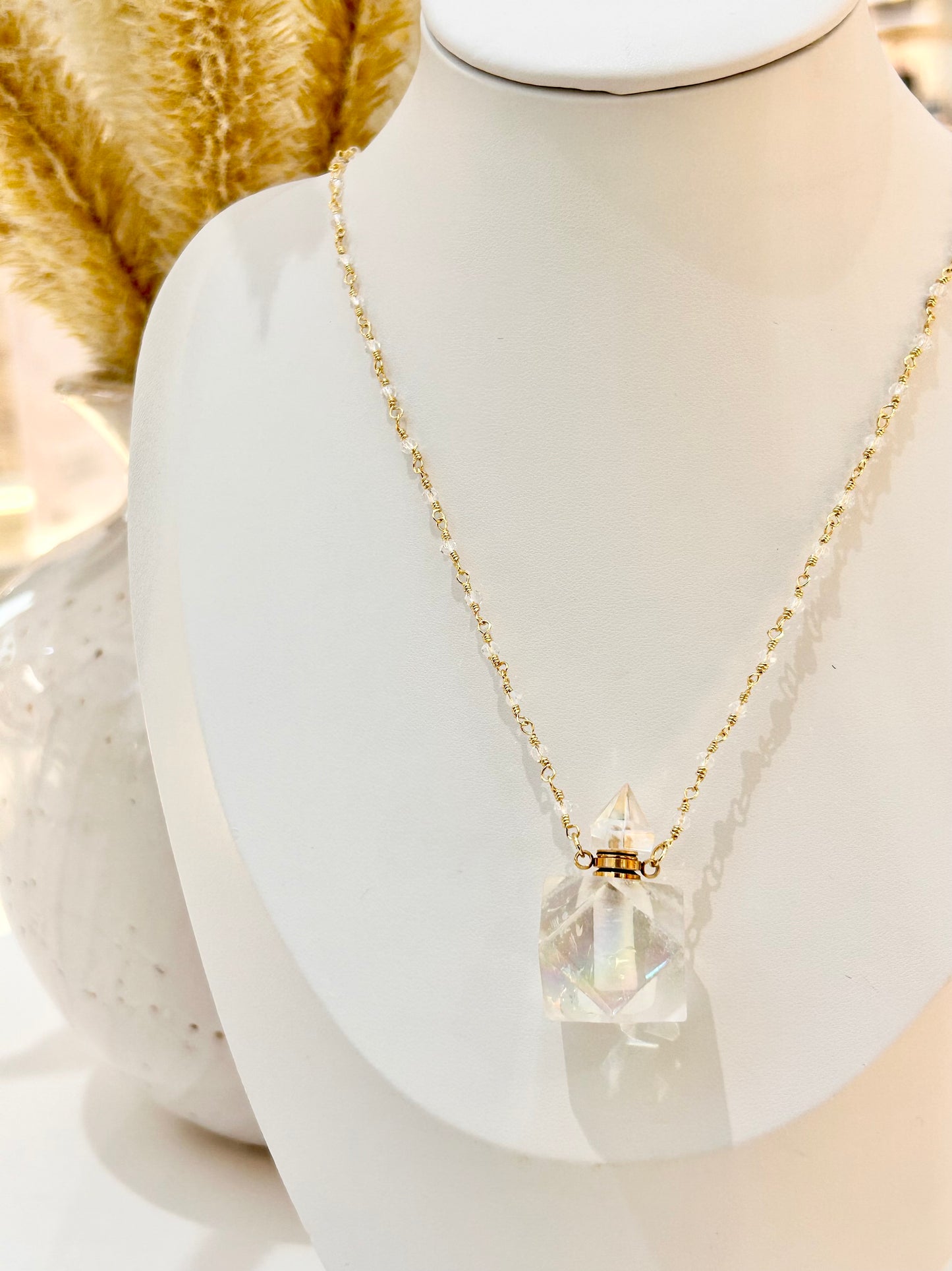 
                  
                    A delicate gold chain necklace with a faceted clear crystal bottle perfume pendant, displayed on a white mannequin bust.
                  
                