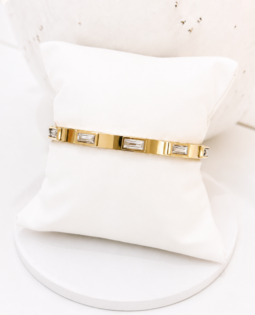 
                  
                    A gold-toned bangle bracelet with rectangular clear stones, displayed on a white cushion stand against a bright white background.
                  
                