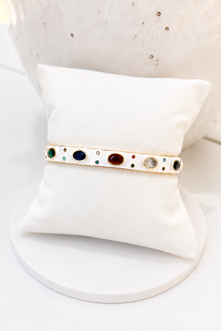 
                  
                    A gold bangle bracelet with colorful gemstone accents, displayed on a white cushion stand.
                  
                