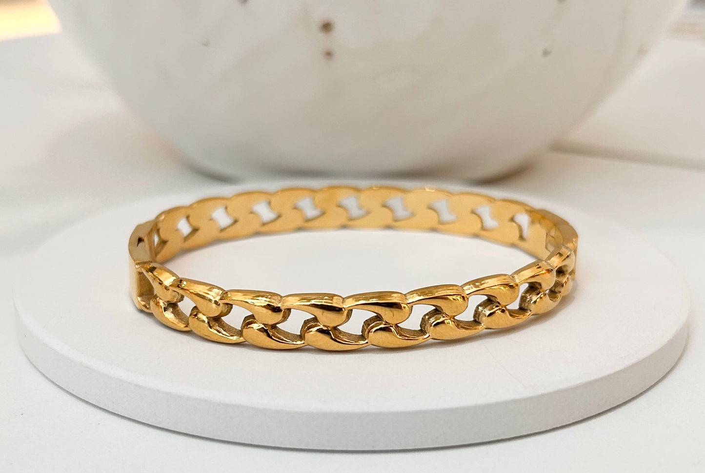 
                  
                    A gold-toned chunky curb chain bangle bracelet displayed on a round white coaster, with a blurred white ceramic bowl in the background.
                  
                