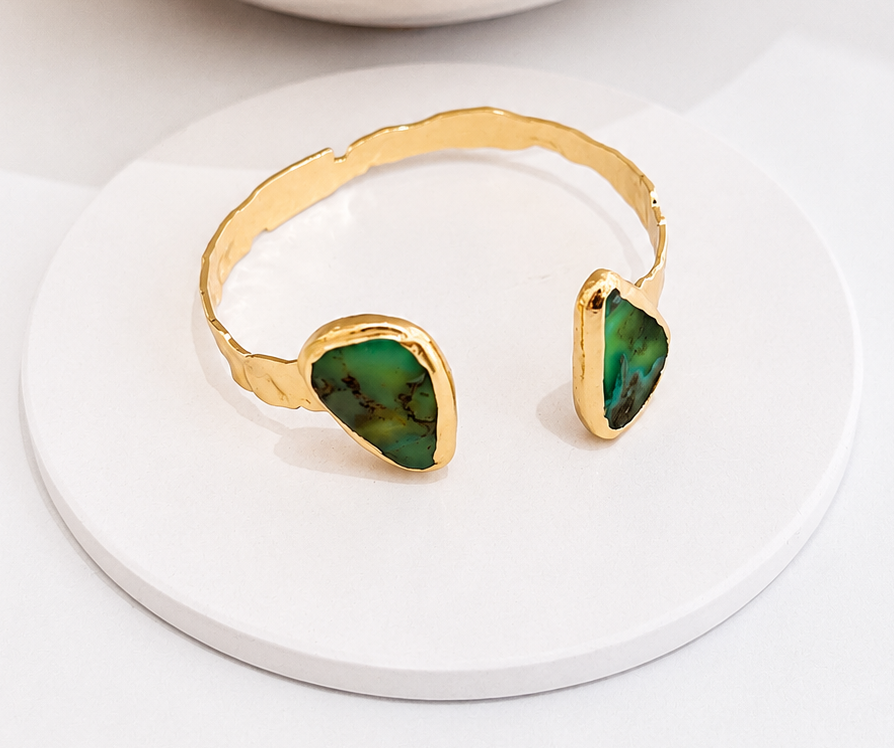 A gold-toned open cuff bracelet with two green chrysoprase stone accents, displayed on a white round coaster with a textured ceramic vase in the background.