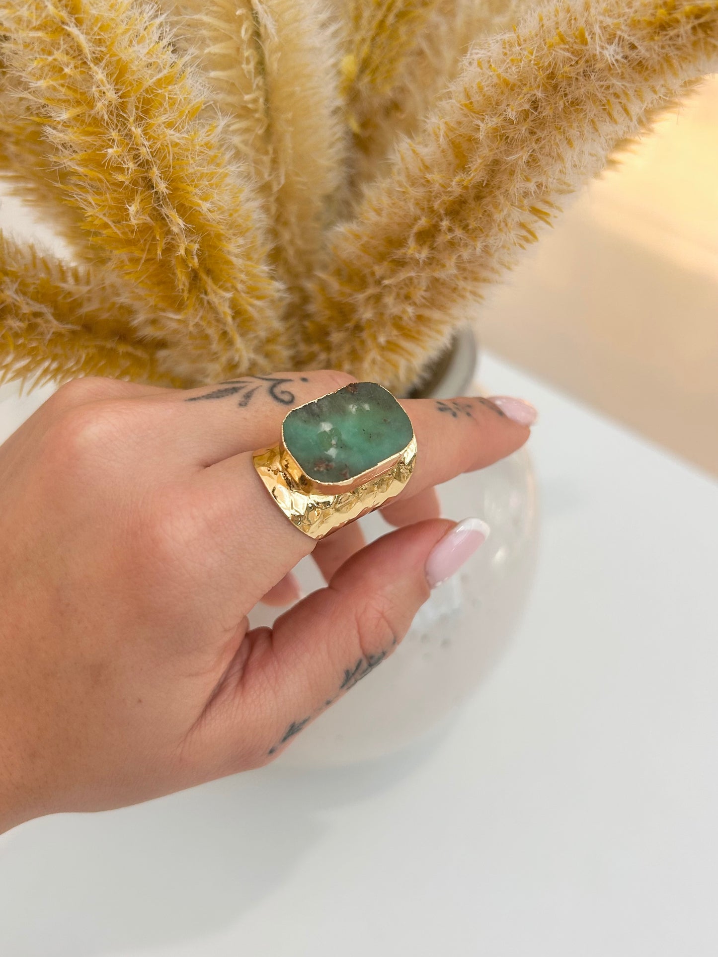 A gold signet ring with a large green Chrysoprase stone worn on a person's hand, with dried pampas grass in a vase in the background.