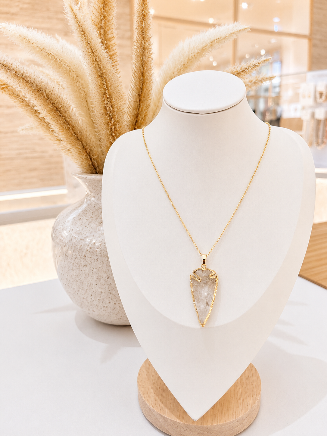 A gold necklace with a teardrop-shaped clear stone pendant displayed on a white jewelry bust, with dried pampas grass in a vase behind it.