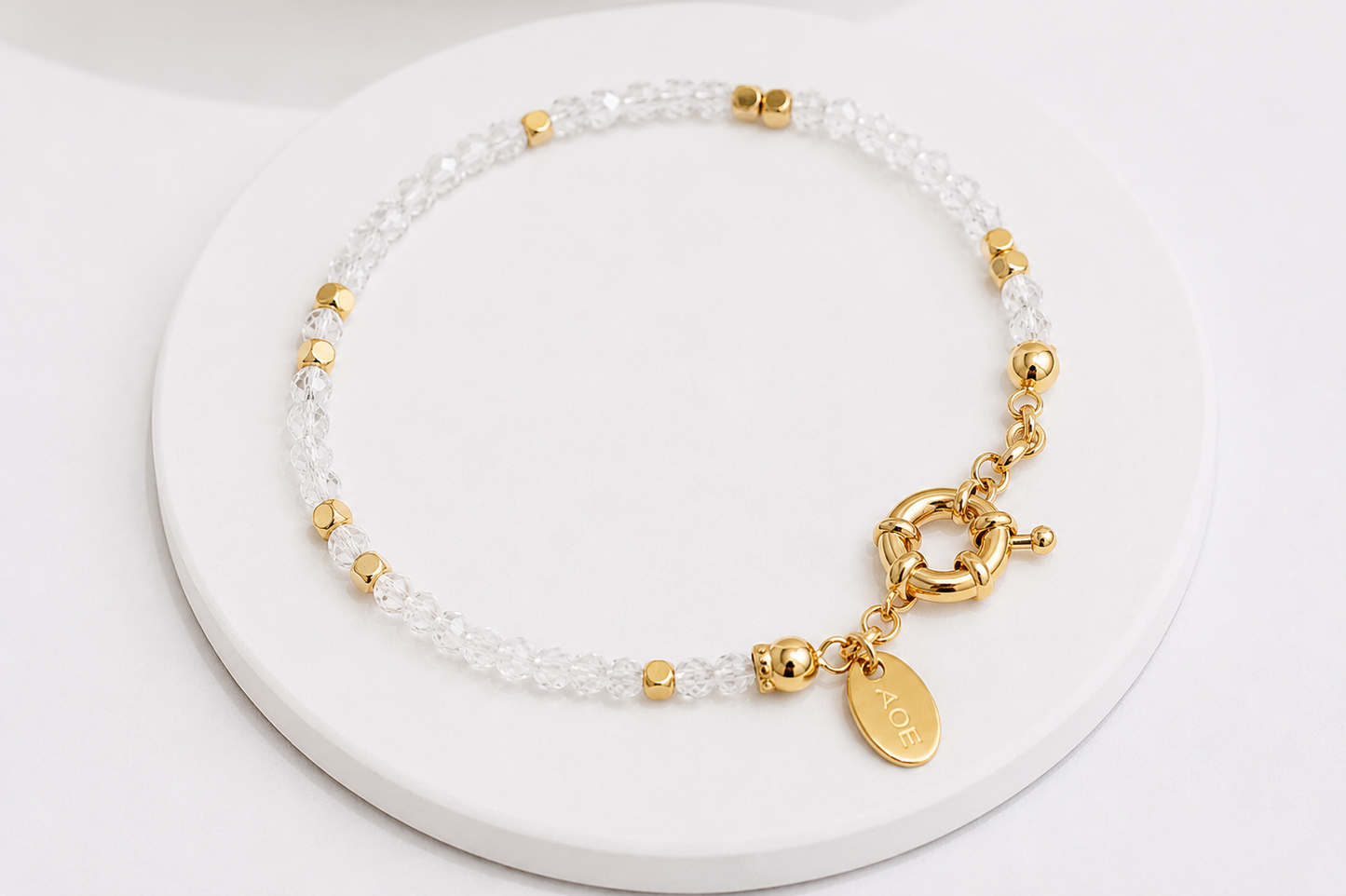 A clear quartz beaded bracelet, small gold spacer beads, and a gold clasp featuring a lobster clasp, chain, and oval charm, displayed on a white round pedestal.