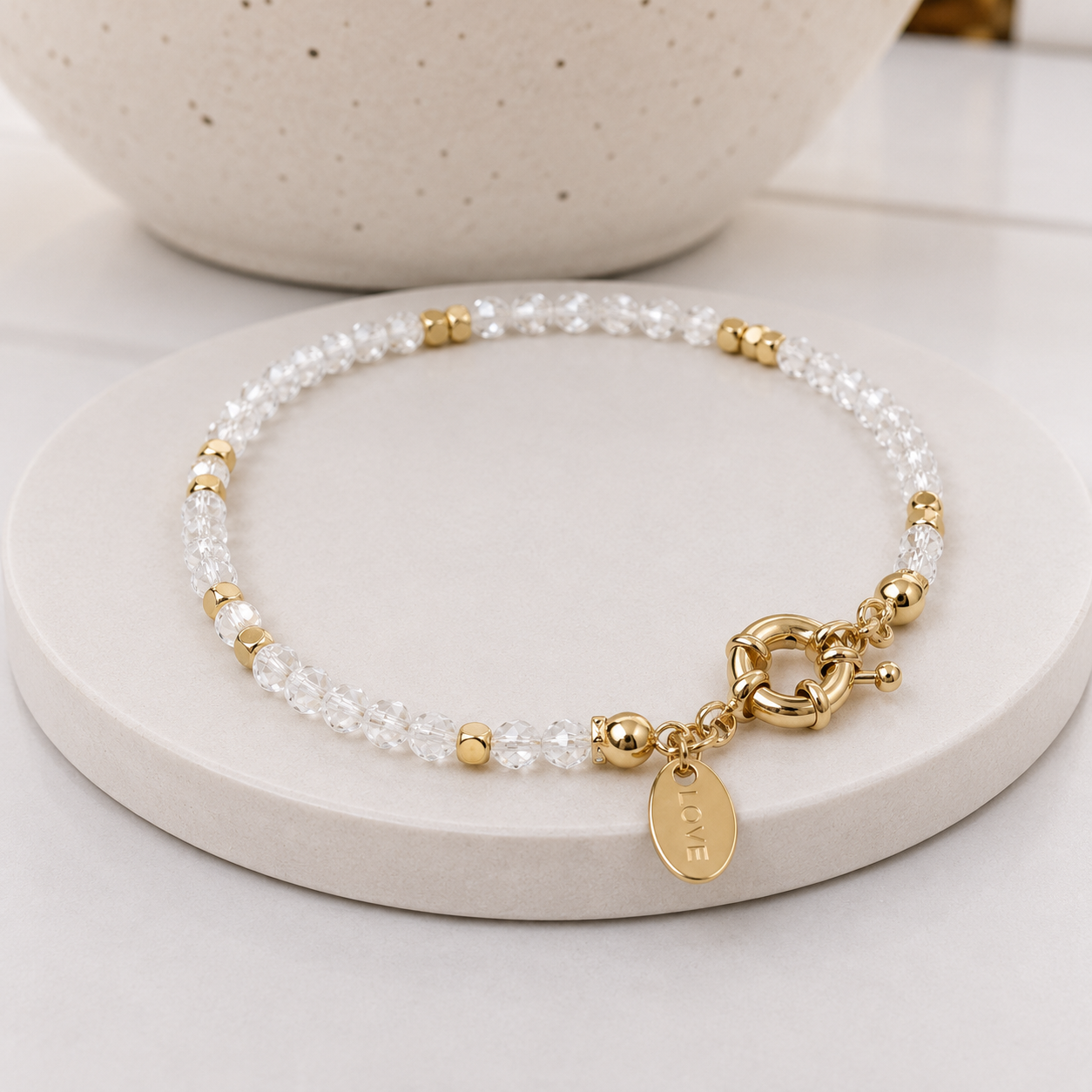 A delicate beaded bracelet with clear quartz beads, small gold spacer beads, and gold-toned hardware, including a toggle clasp and a round charm engraved with 'LOVE'. The bracelet is displayed on a round beige stone stand with a ceramic bowl in the background.