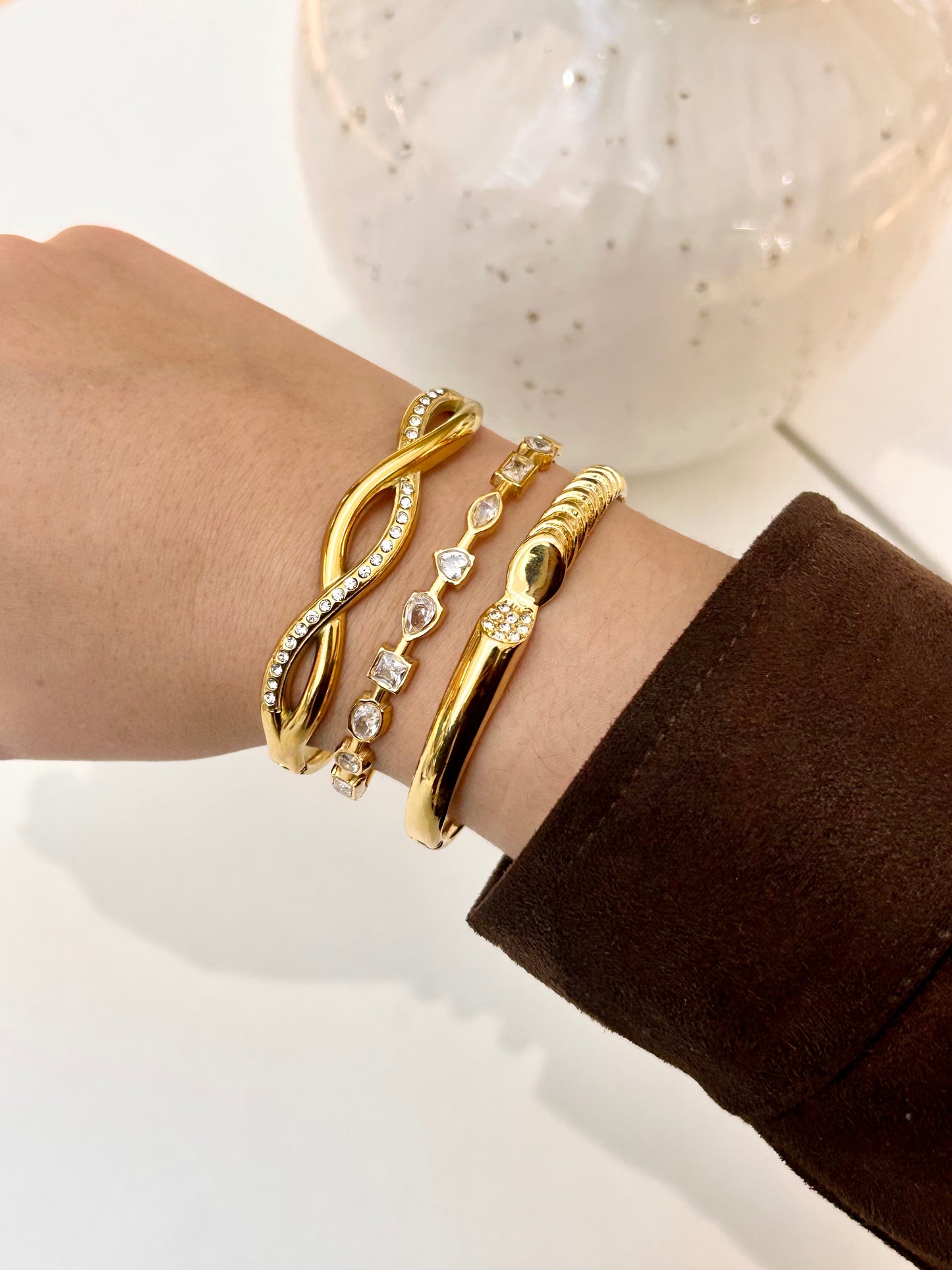 A close-up of a person's wrist wearing four gold-toned bracelets, including an infinity bracelet with clear stones, a beaded chain bracelet, and two polished gold bands, with a white speckled vase blurred in the background.