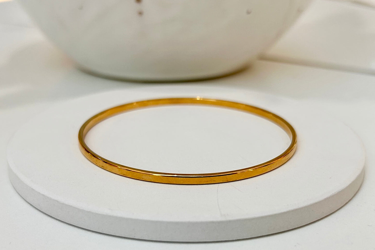 A thin, polished gold bangle bracelet displayed on a round white coaster, with a blurred white ceramic bowl in the background.