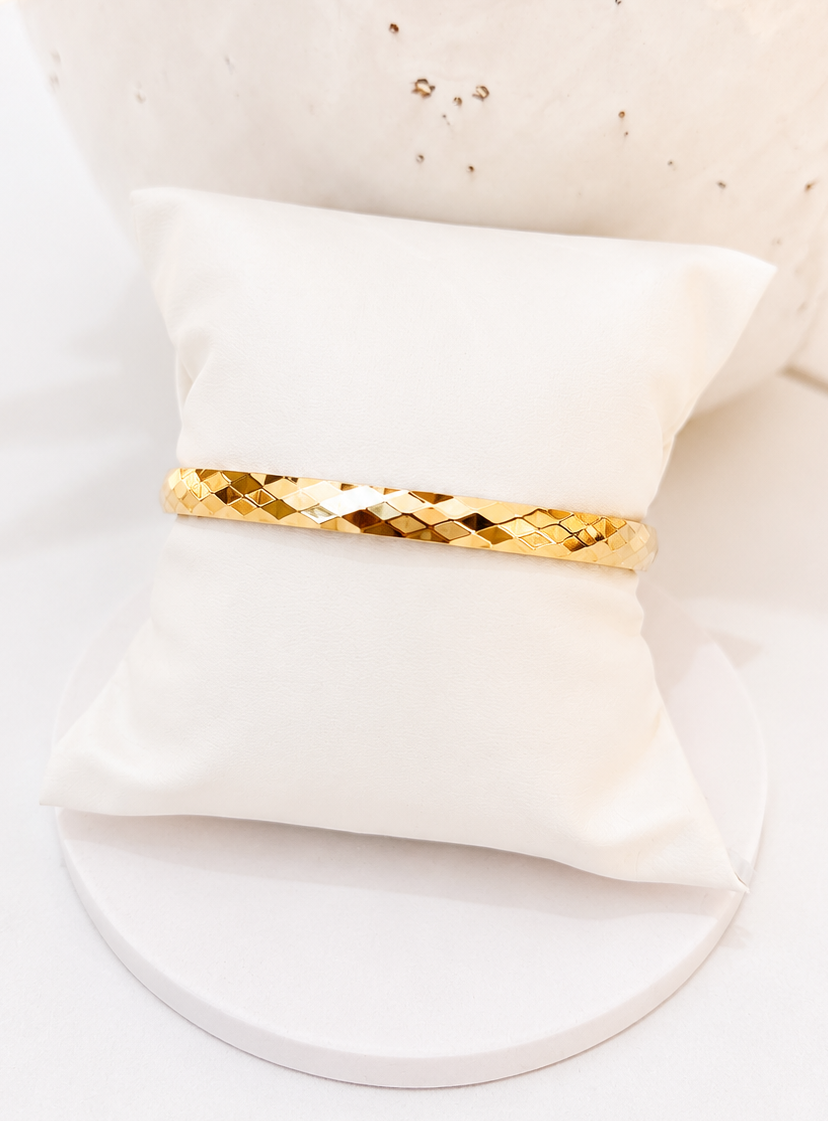 
                  
                    A gold diamond-patterned bangle bracelet displayed on a small white fabric pillow, set against a bright white background with a textured white vase in the background.
                  
                