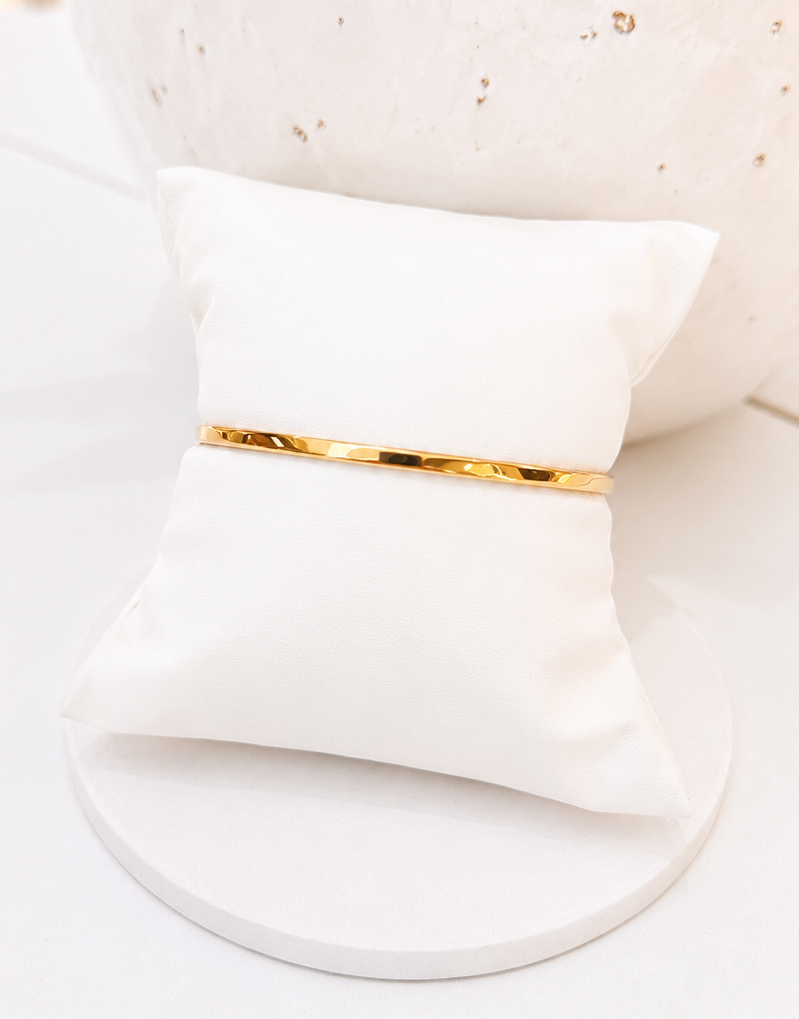 
                  
                    A slim gold bangle bracelet displayed on a small white fabric jewelry pillow, set against a clean white background with a decorative white vase in the background.
                  
                