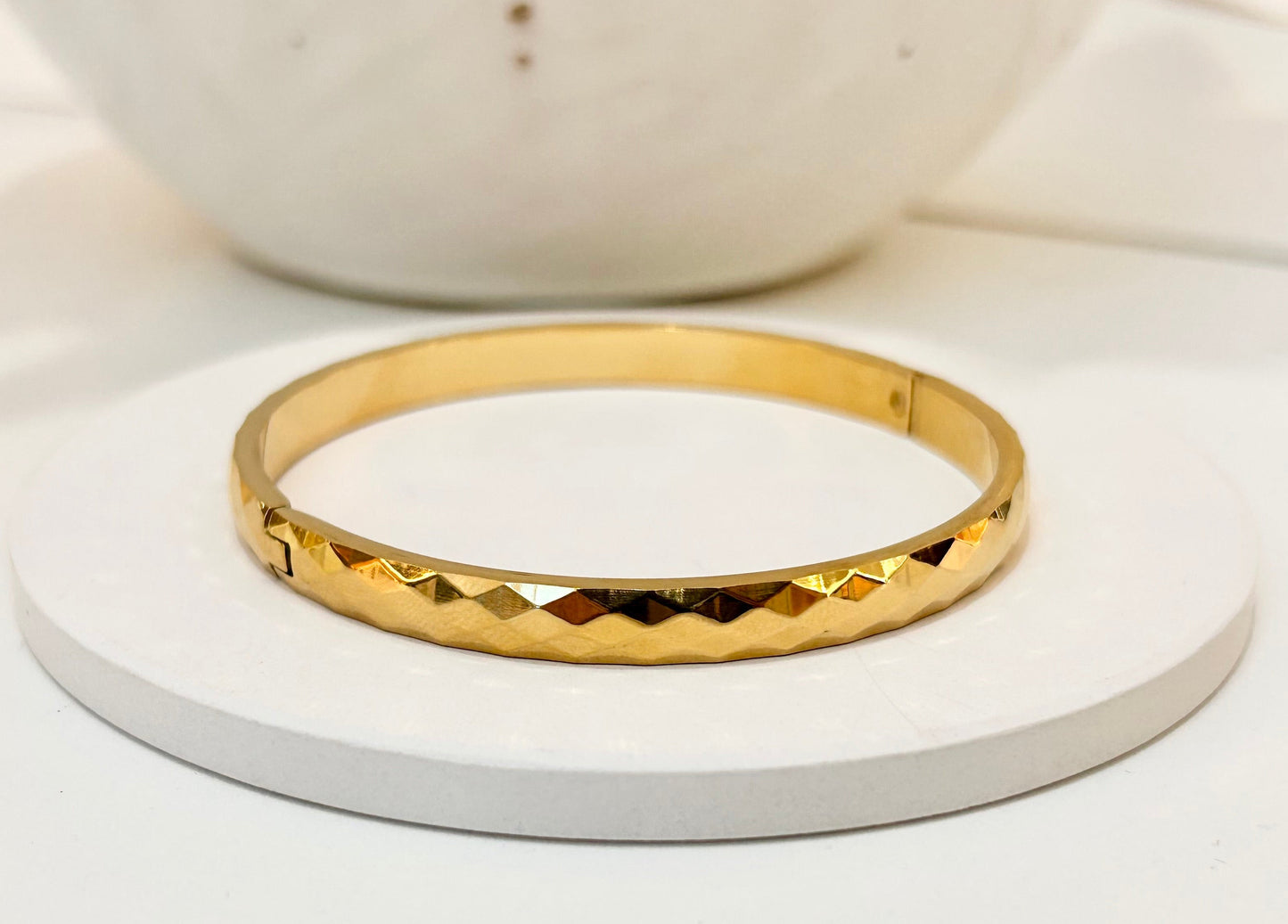 A gold bangle bracelet with a textured, diamond-cut pattern, displayed on a round white coaster.