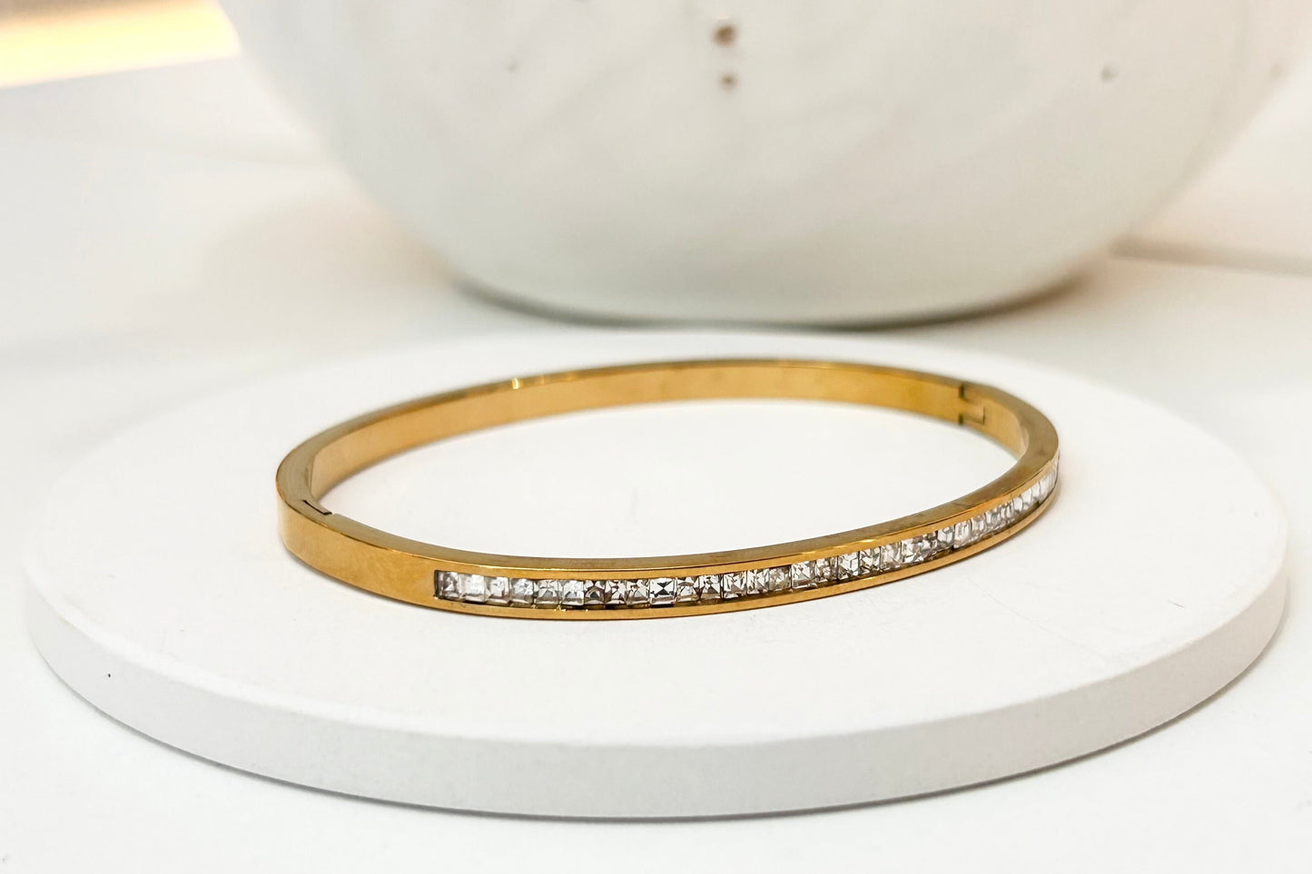 
                  
                    A gold bangle bracelet with a row of small clear stones, displayed on a round white coaster with a white ceramic vase in the blurred background.
                  
                