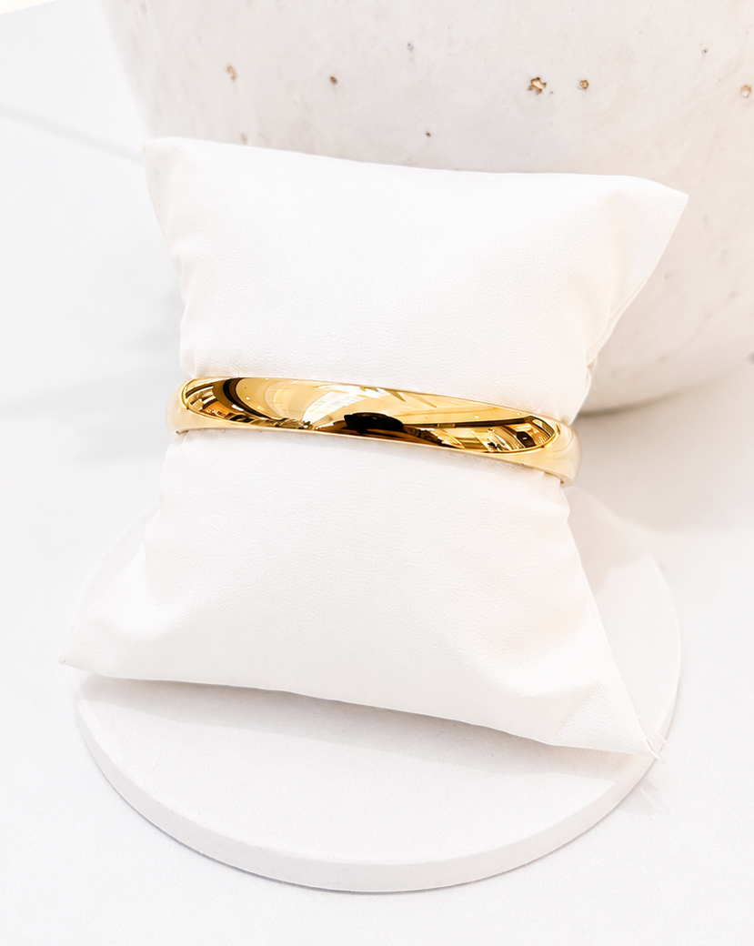 
                  
                    A polished gold band ring displayed on a small white fabric ring holder, set against a bright white background with a decorative white textured vase.
                  
                