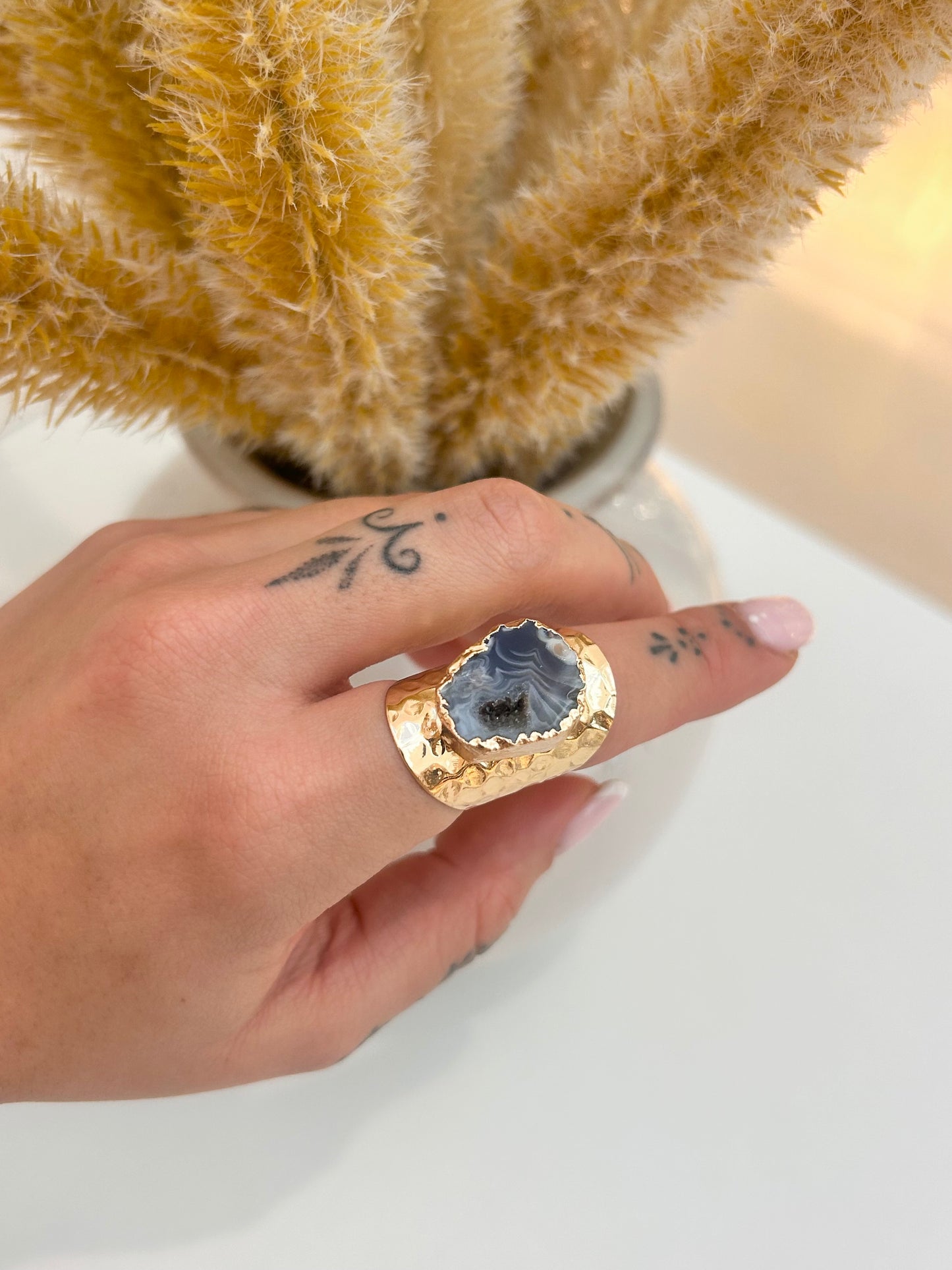 A close-up of a hand wearing a chunky gold ring with a textured, hammered finish and a large, irregular dark gray agate stone. The hand is resting on a white surface with dried pampas grass in the background.