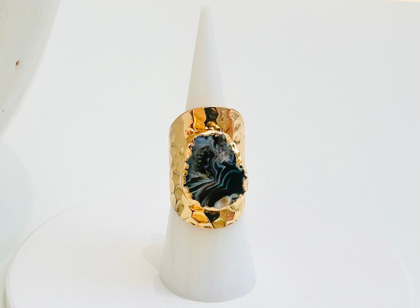 
                  
                    A gold ring with a large, irregularly shaped black and gray agate slice centerpiece, displayed on a white cone stand against a white background.
                  
                