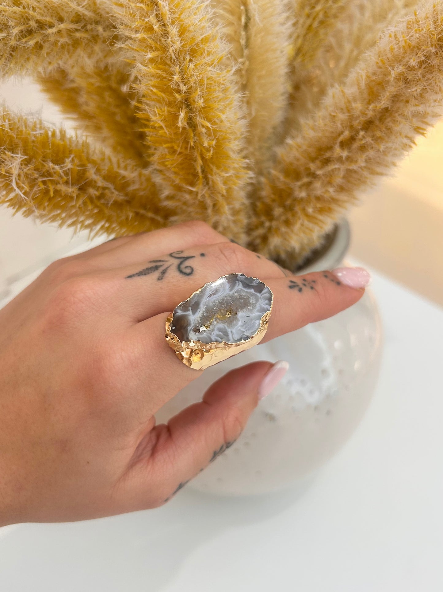 A gold ring featuring a large, raw gray geode adjustable ring, shown worn on a hand with black tattoos, set against a background of dried pampas grass in a white vase.