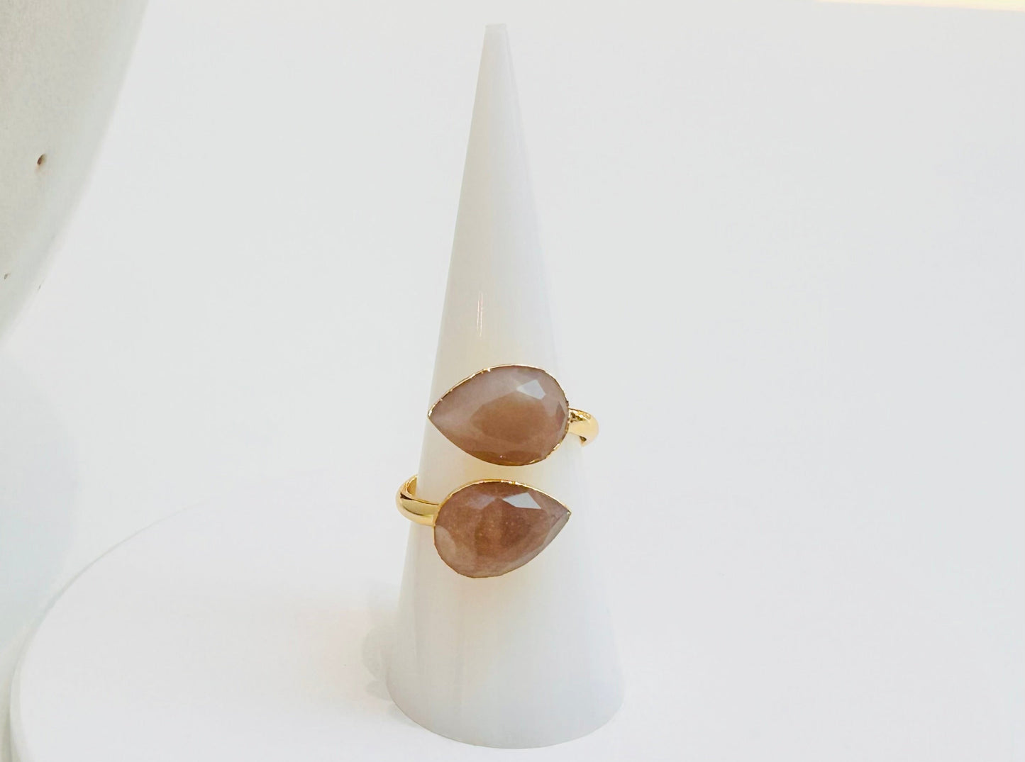 
                  
                    A gold ring with two teardrop-shaped sunstone gemstones displayed on a white cone ring stand against a white background.
                  
                