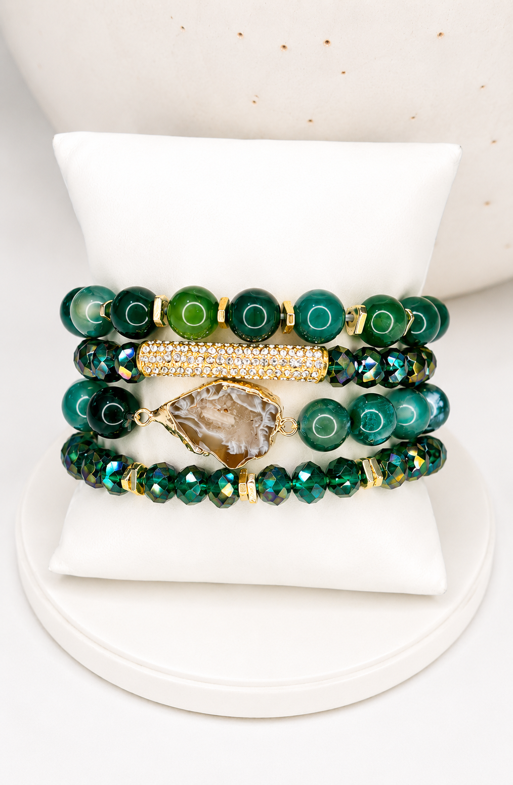 A stack of four beaded  natural stone stretch bracelets displayed on a white jewelry stand. The bracelets feature green beads, iridescent dark green beads, a gold beaded accent, and a central natural druzy stone charm.