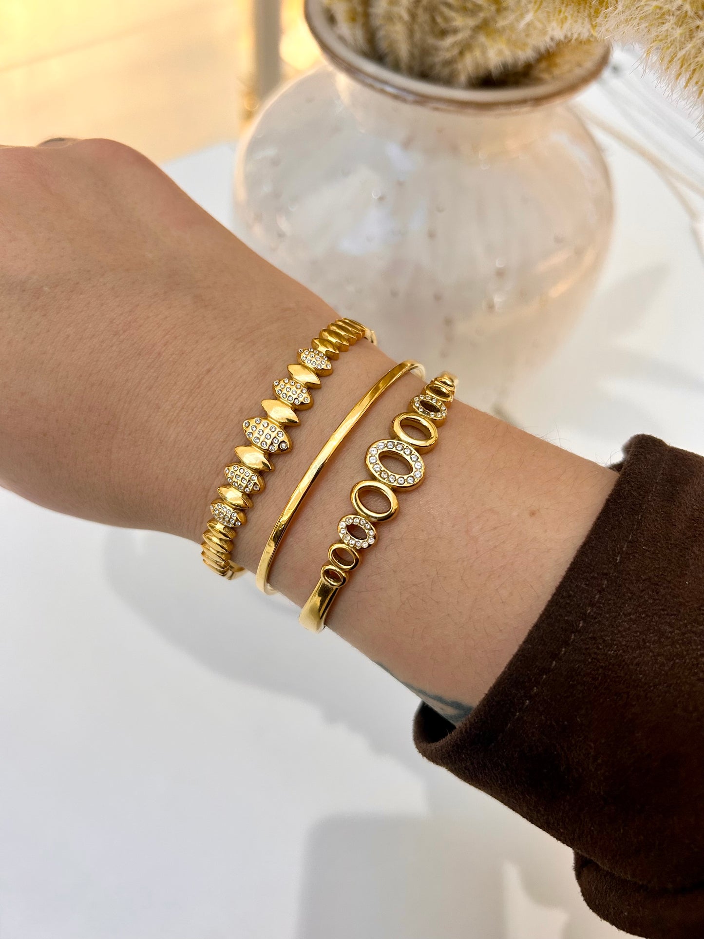 A close-up of a person's wrist wearing three gold bracelets, including one with textured spikes, one with a smooth bar, and one with oval links set with small clear stones. A brown sleeve is visible, with a blurred decorative vase in the background.