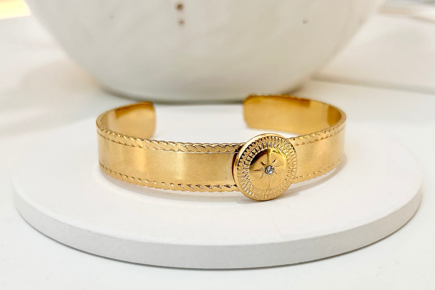 A gold cuff bracelet with textured rope-like detailing and a round decorative clasp, displayed on a white round pedestal.