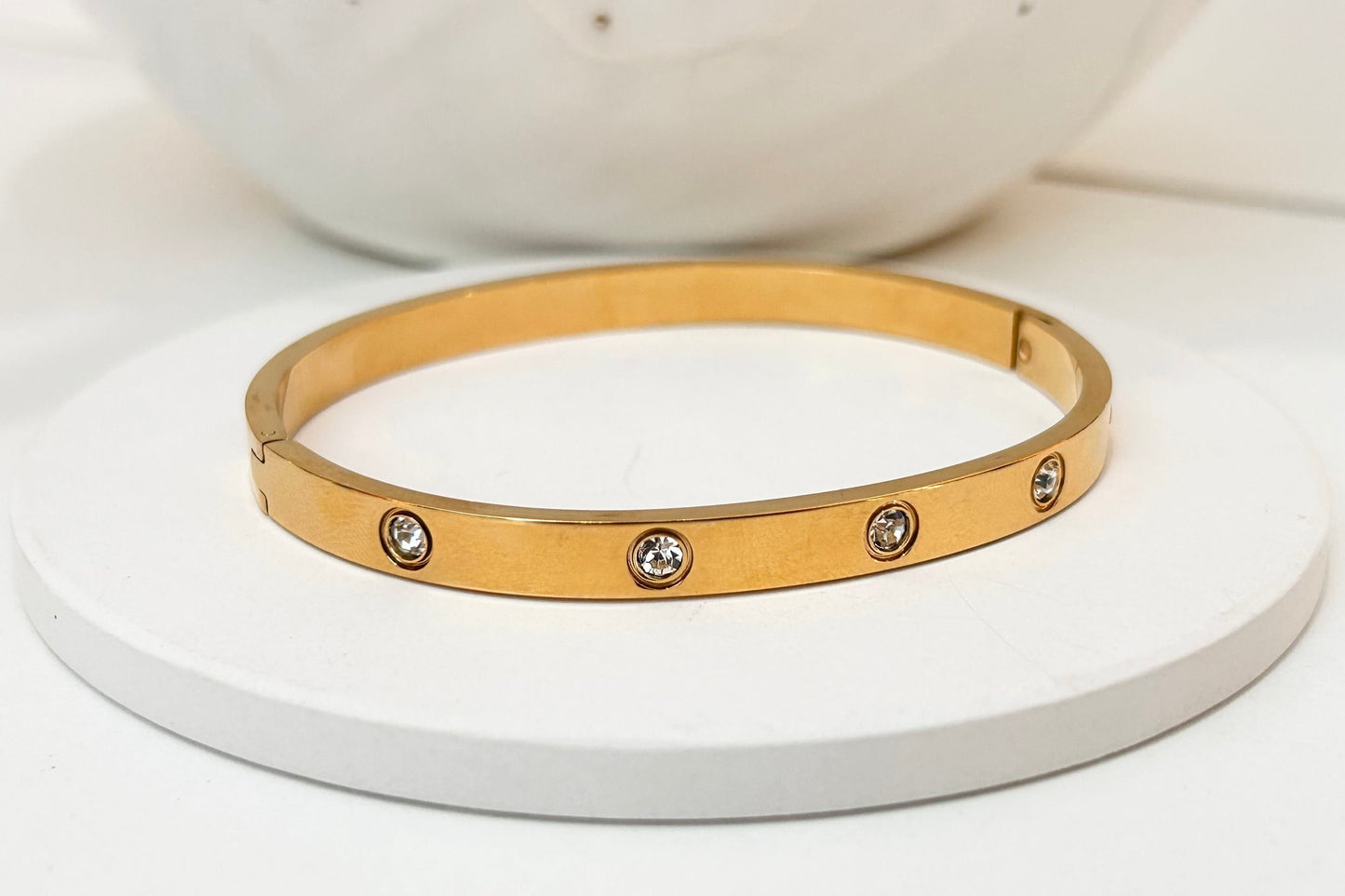 A gold bangle bracelet with five small bezel-set clear stones, displayed on a round white pedestal.