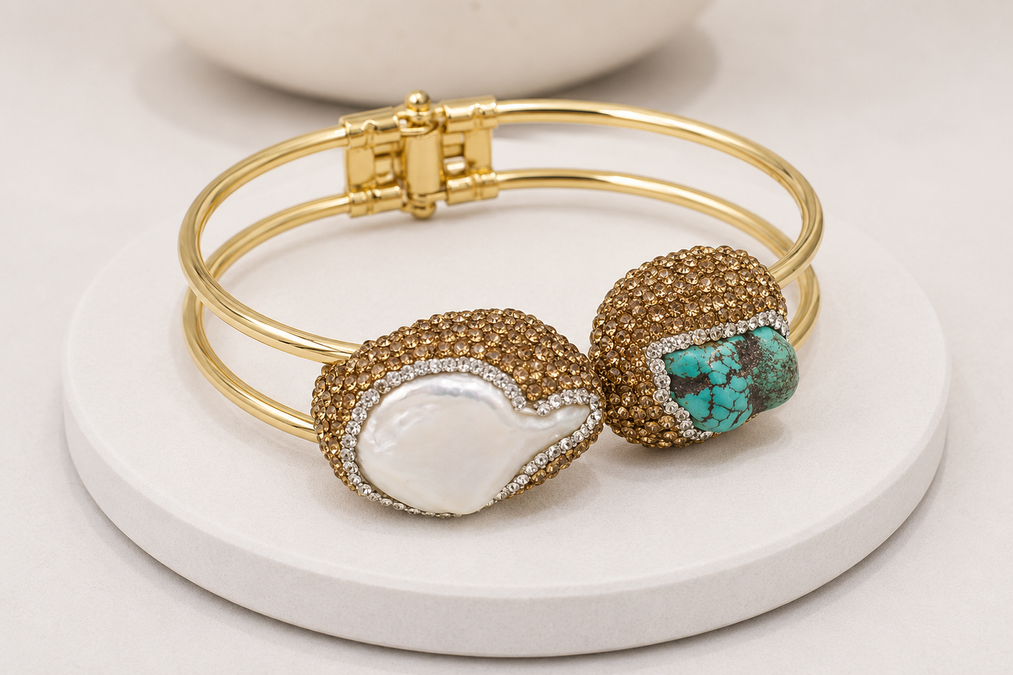 A gold-plated bangle bracelet with two decorative oval charms, one featuring a white mother-of-pearl inlay and the other featuring a turquoise stone surrounded by pavé crystals, displayed on a white round pedestal.