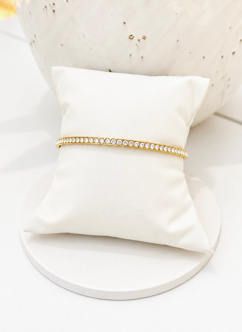 
                  
                    A delicate gold tennis bracelet with a row of clear cubic zirconia stones, displayed on a white satin pillow.
                  
                