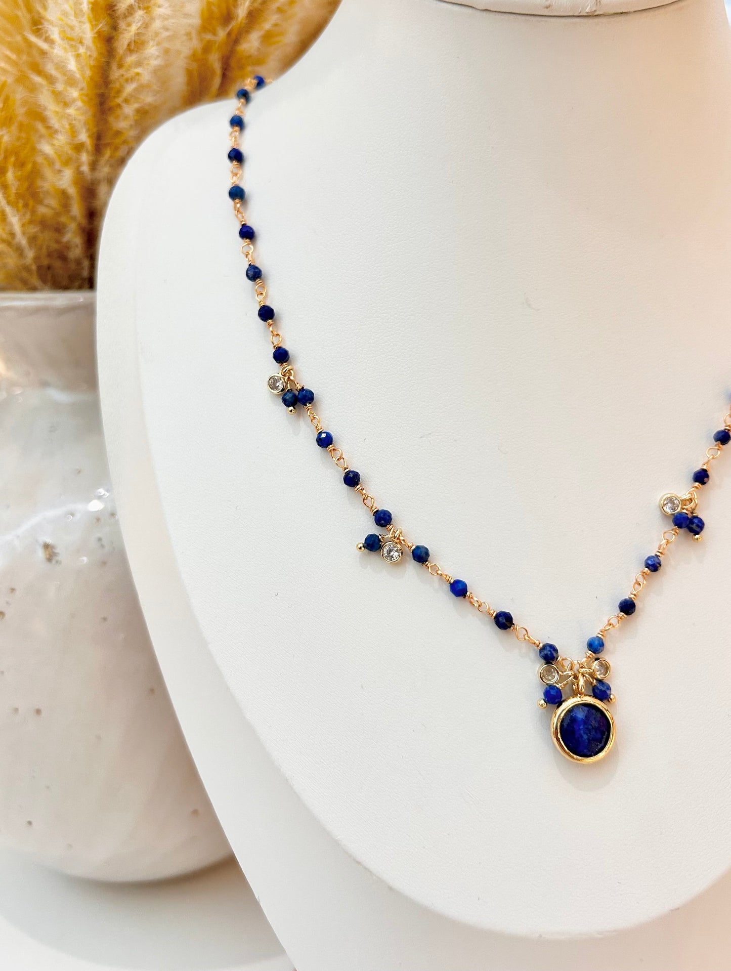 
                  
                    A beaded necklace with deep blue lapis lazuli beads, small gold-toned floral accents, and a round blue stone pendant in a gold bezel, displayed on a white jewellery bust.
                  
                