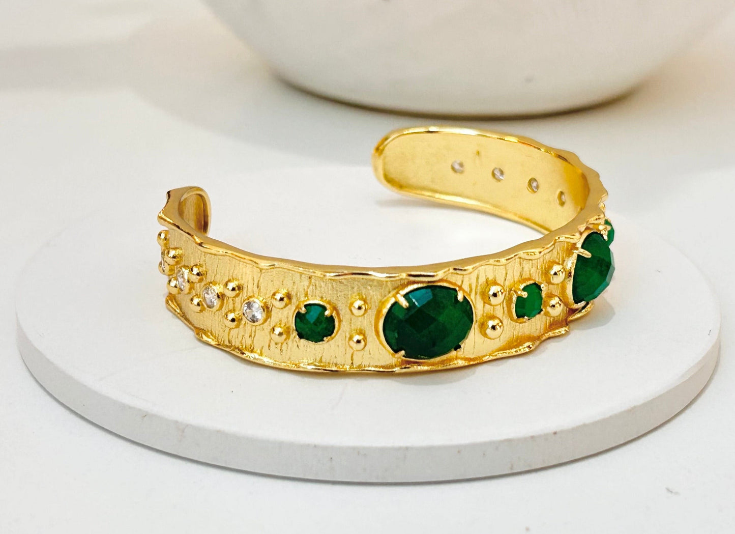 A gold cuff bracelet with green jade gemstone accents, displayed on a round white coaster with a white speckled bowl in the background.