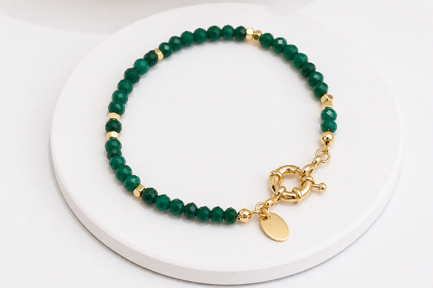 A beaded bracelet with faceted dark green jade beads, small gold spacer beads, and a gold toggle clasp with a round tag, displayed on a white circular pedestal.