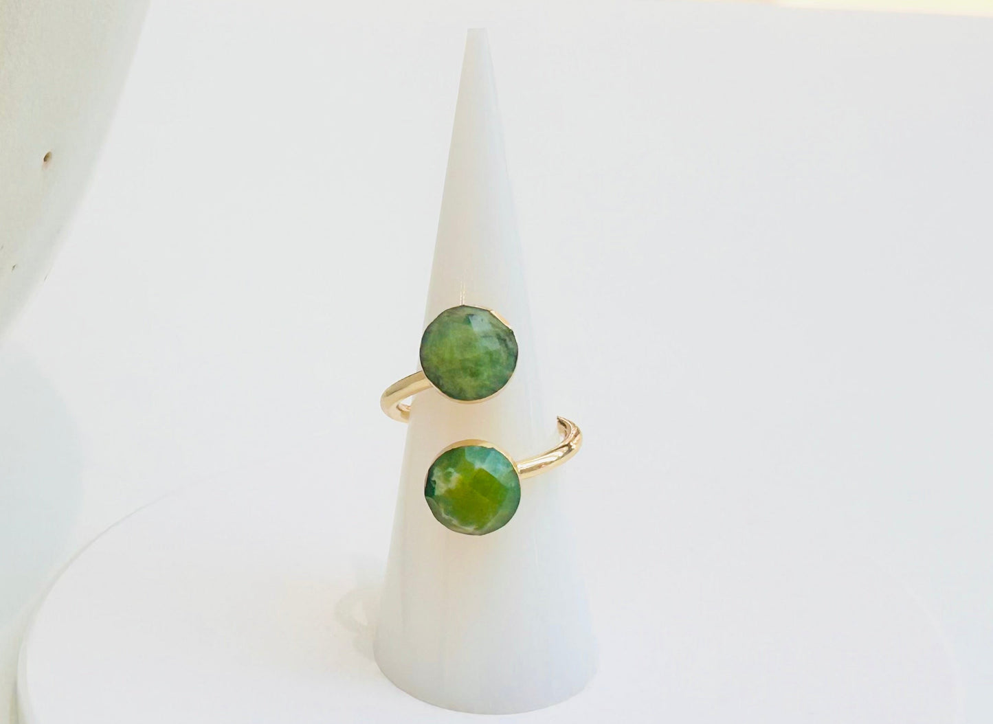 
                  
                    A gold ring with two round green jade gemstones displayed on a white cone ring stand, set against a clean white background with a soft-focus ceramic object on the left.
                  
                