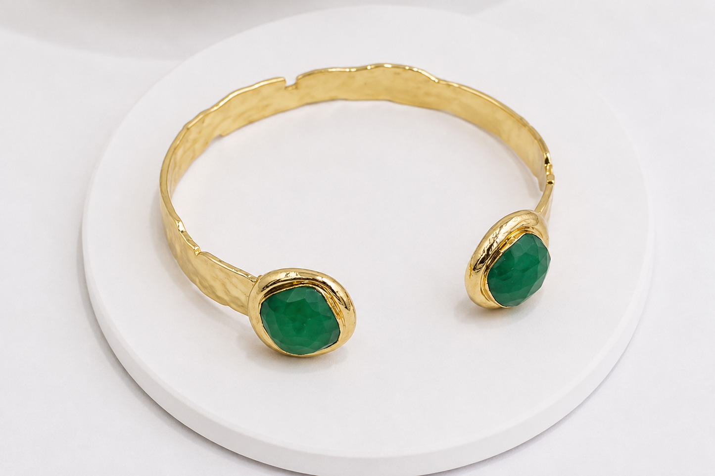A gold-toned open cuff bracelet with two oval green jade gemstone accents, displayed on a round white pedestal against a white background.