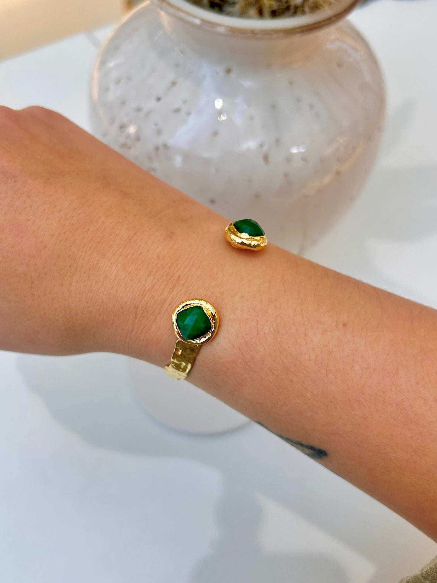 A close-up of a person's wrist wearing gold cuffs, featuring a round green jade stone, with a white speckled vase blurred in the background.