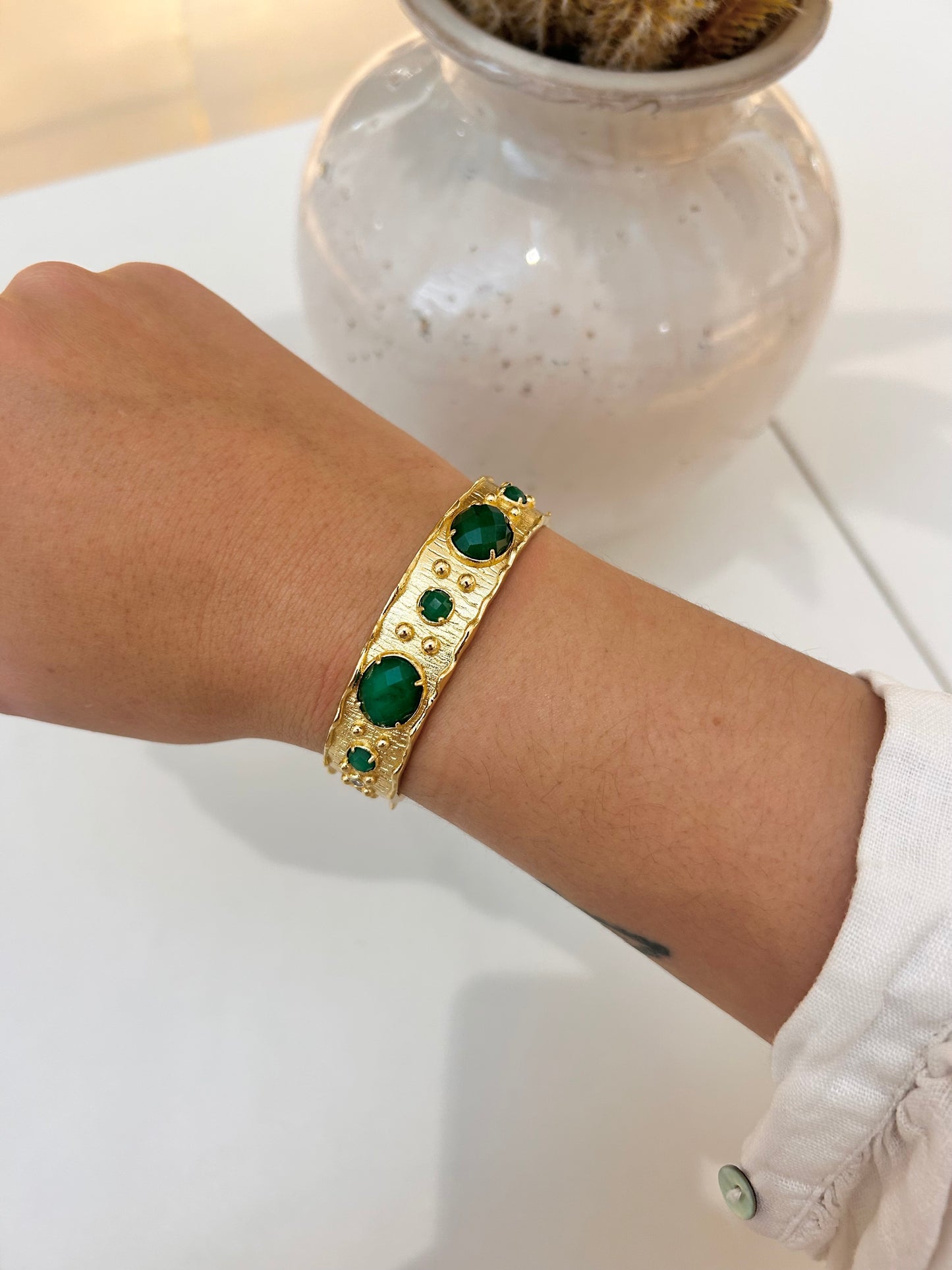 A gold-toned cuff bracelet with green jade gemstone accents worn on a person's wrist, shown against a white tabletop with a blurred decorative vase in the background.